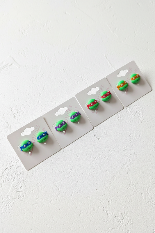 All four TMNT Ninja Turtle stud earring variants on white display cards — Leonardo blue, Donatello purple, Raphael red, Michelangelo orange — styled in a clean Pinterest flat lay